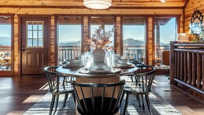 A wooden interior with a round dining table set for six, surrounded by chairs, and a large window offering a view of the mountains.