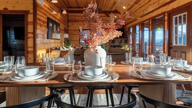 A wooden cabin with a dining area set for a meal, featuring a large vase with flowers, wooden walls, and large windows.