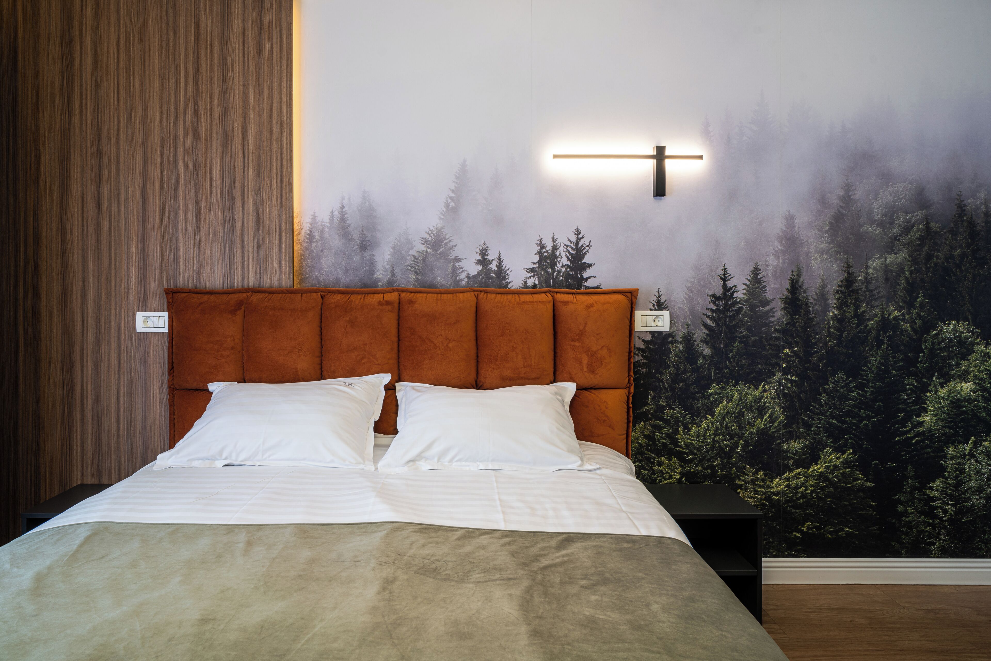 A bedroom with a large bed, a headboard, a nightstand, and a wall mural of a forest.