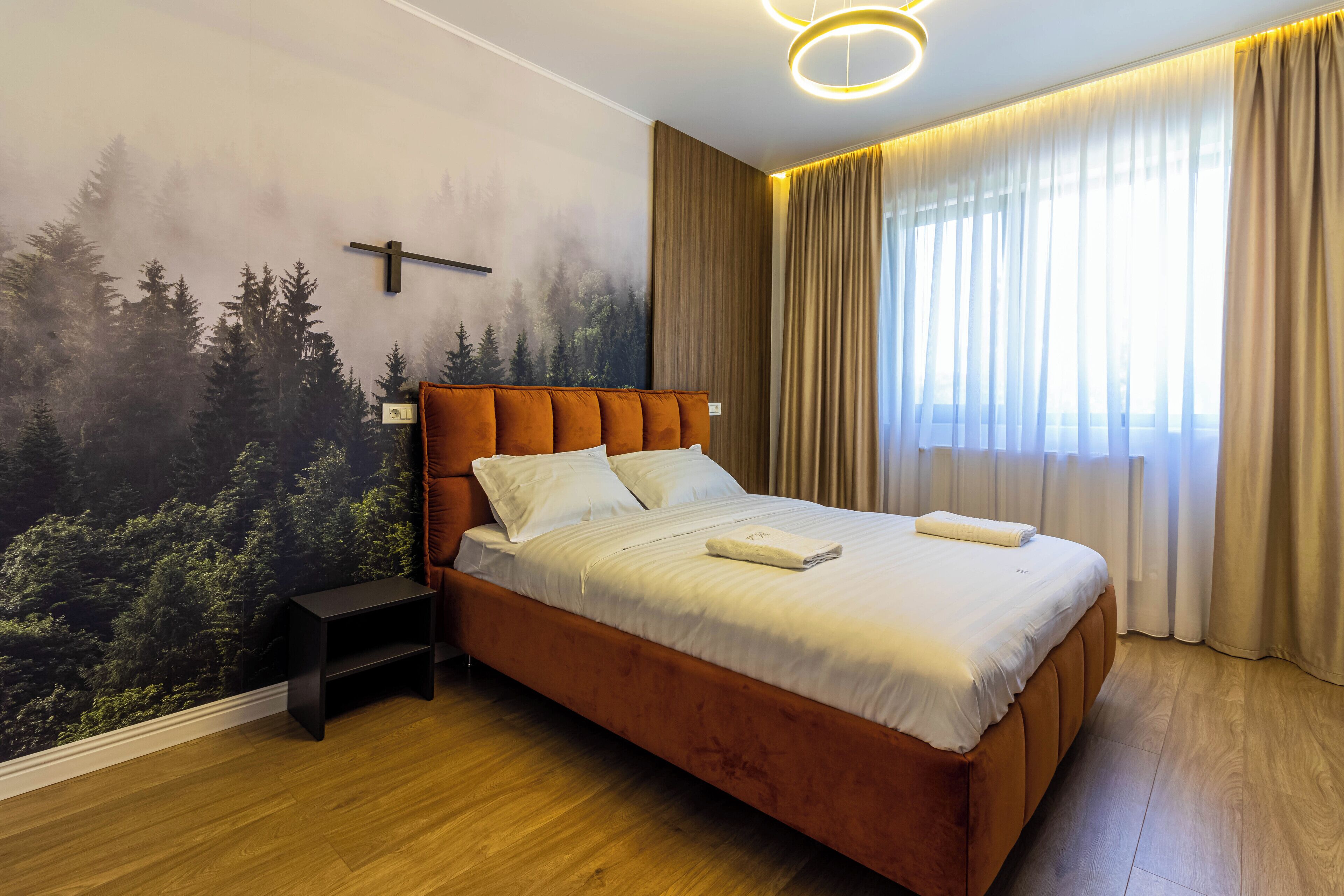 A bedroom with a large bed, a nightstand, a wall mural, and a window with curtains.