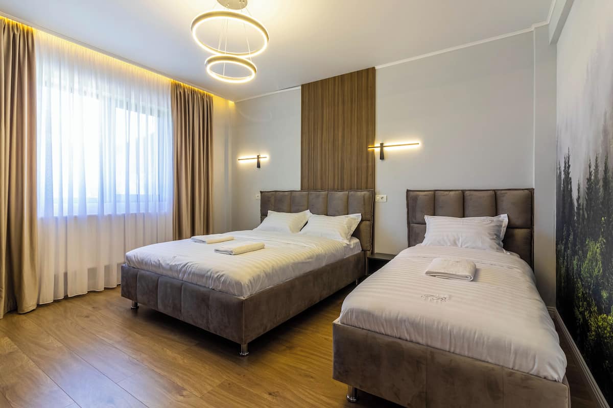 A hotel room with two beds, wooden flooring, a large window with curtains, wall-mounted lights, and a painting of trees.
