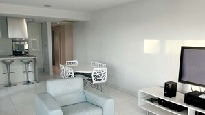 A modern living room with a white sofa, a coffee table, and a TV stand with a flat-screen TV. There is a kitchen area in the background with a white countertop and a stainless steel range hood. - Modern seafront apartment (Limassol)