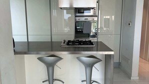 A modern kitchen with a stainless steel range hood, double oven, and two bar stools. - Modern seafront apartment (Limassol)