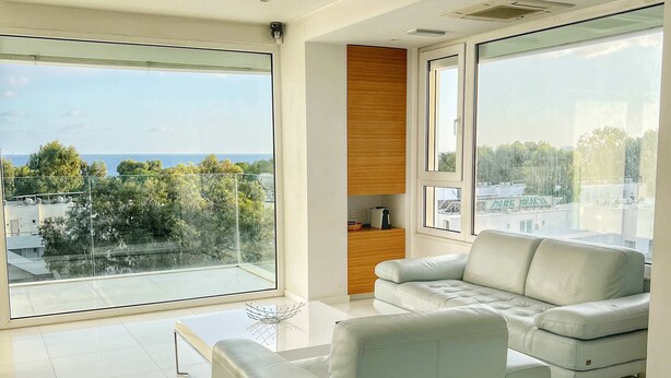A modern living room with a large window, a white sofa, a coffee table, and a dining area with chairs. - Modern seafront apartment (Limassol)