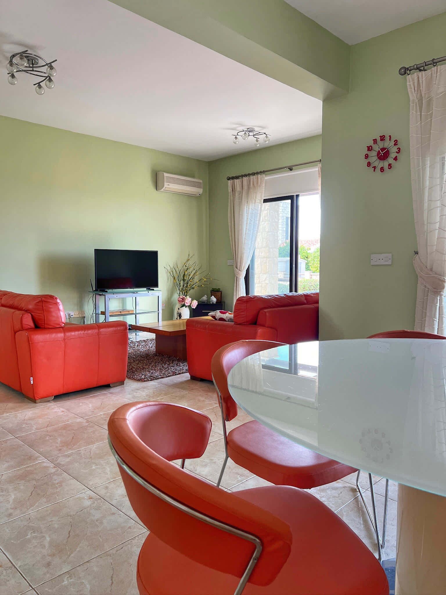 A living room with a red sofa set, a glass dining table with chairs, a flat-screen TV, and a large window with curtains.
