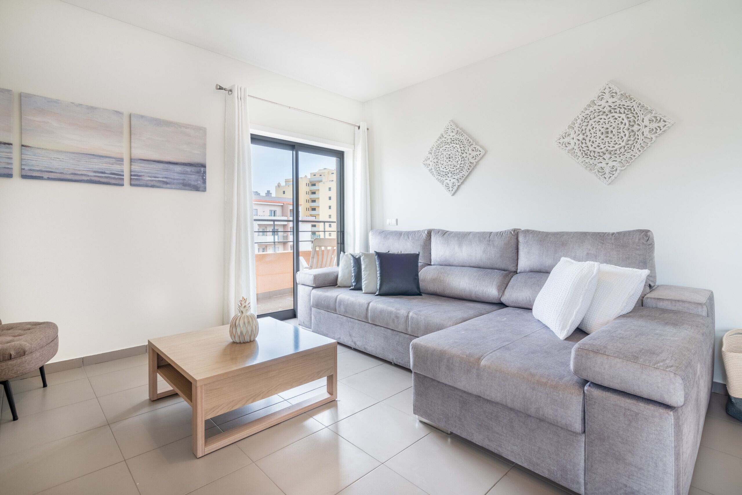 A modern living room with a grey sofa, a wooden coffee table, and a view of buildings through a large window.