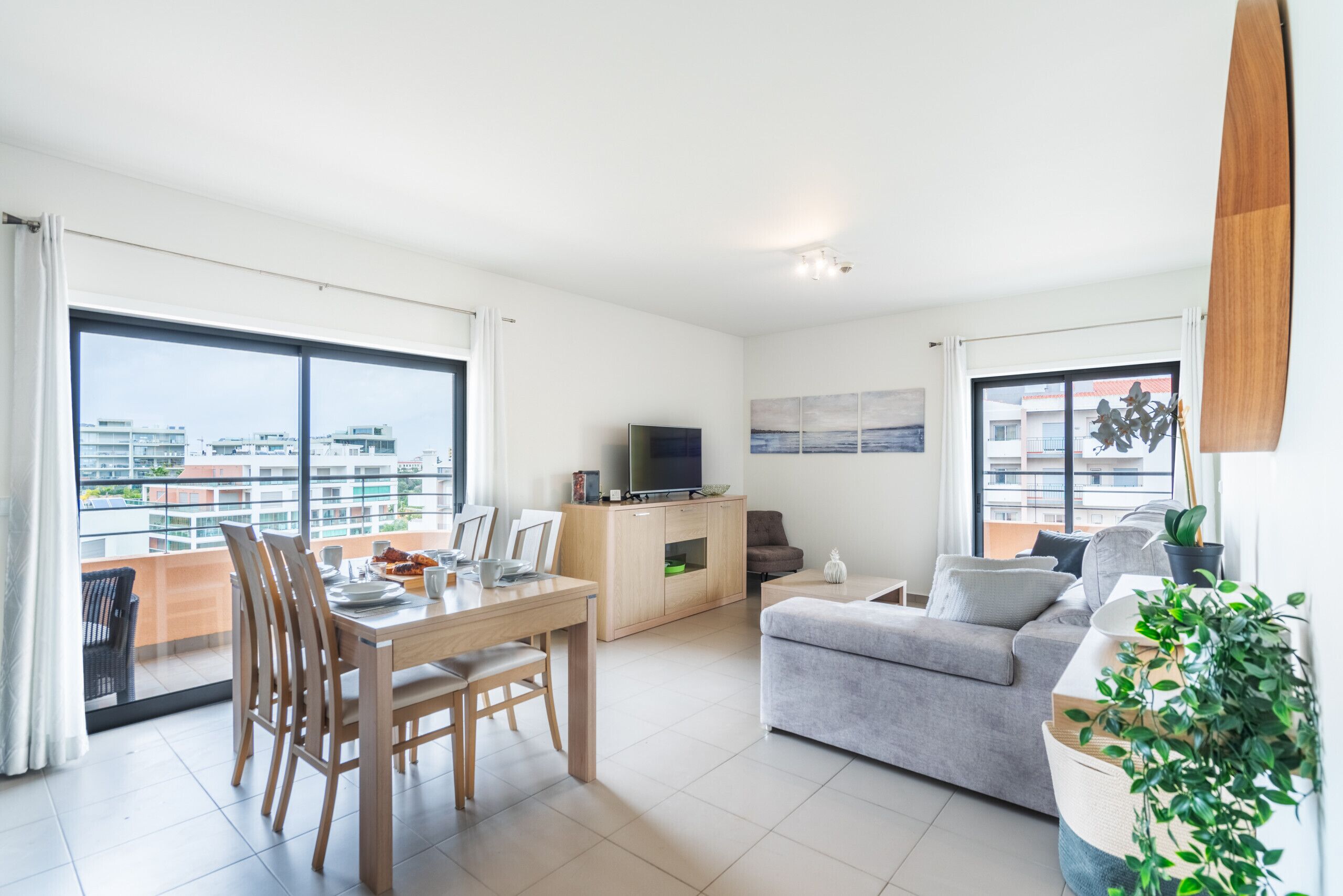 Un salon moderne comprenant un coin repas, un canapé gris, un téléviseur et un balcon offrant une vue sur la ville.