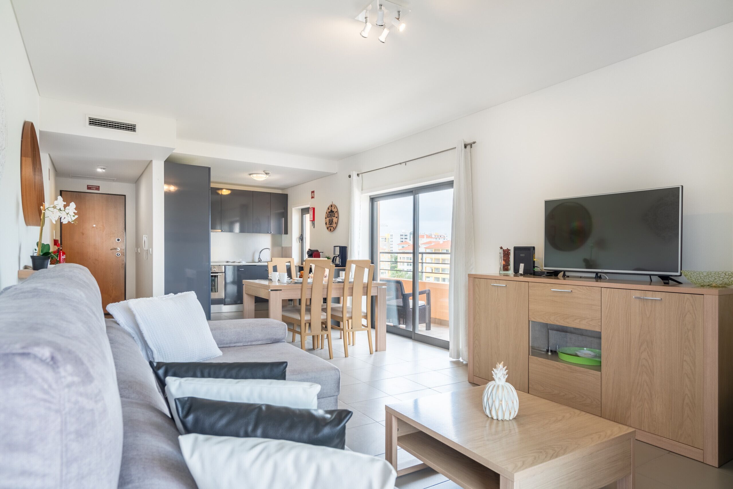 Ein modernes Wohnzimmer mit einer Couch, einem Couchtisch und einem Fernseher. Es gibt eine Küchenzeile mit einem Esstisch und Stühlen.