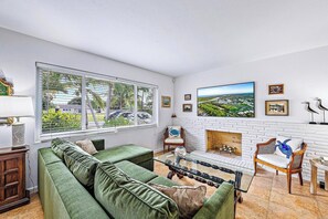 A living room with a green sofa, a glass coffee table, a fireplace, and a large window with a view of greenery. - Casa Balsinde - Delight in a Private Pool in the Southern End of West Palm (West Palm Beach)