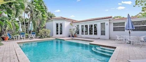 A swimming pool with a tiled deck, surrounded by white buildings with windows and doors, and outdoor furniture.