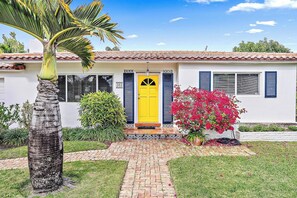 Una casa con una puerta amarilla, un sendero de ladrillo y una palmera.