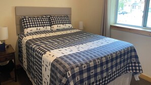 Ein Schlafzimmer mit kariertem Bettbezug, einem Holzkopfteil, Nachttischlampen und einem Fenster mit Blick auf Gebäude.