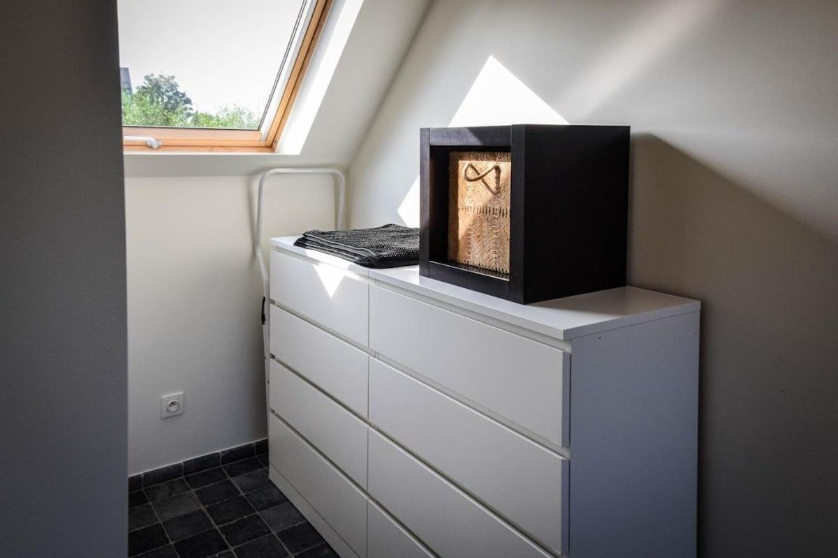 Un coin de pièce avec une commode blanche, un tableau encadré et une lucarne.