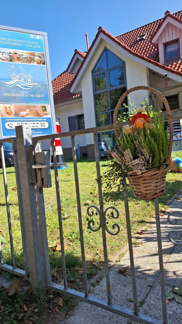 En grind av metall med en korg full av blommor, ett hus med ett rött tegeltak och en skylt i bakgrunden.