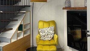 Uma sala de estar com escada, poltrona amarela, relógio redondo na parede e lareira.
