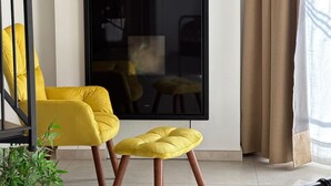 Uma sala de estar moderna com poltrona e banquinho amarelos, uma TV grande com moldura preta e uma estante de madeira.