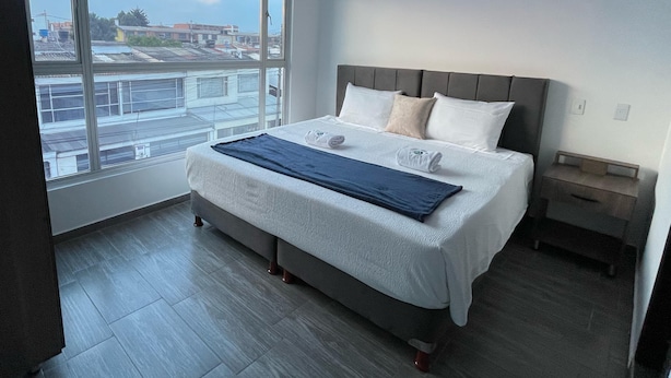 A modern bedroom with a large window, a bed with a grey headboard, and a wooden floor. - Outlanders House (Bogota)
