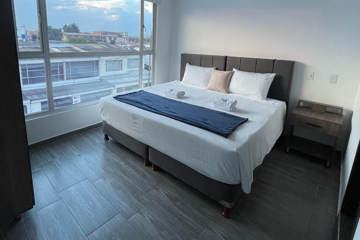 Un dormitorio moderno con un ventanal grande, una cama con cabecero gris y suelo de madera.