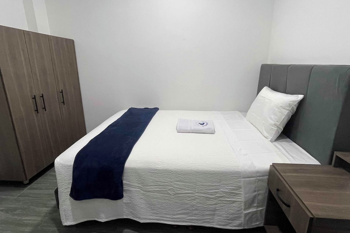Una cama bien hecha con edredón blanco y manta azul, cabecera gris y armario de madera.