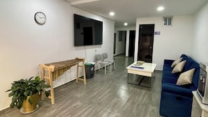A modern living room with a blue sofa, a wooden bench, a TV mounted on the wall, a clock, and a small potted plant. - Outlanders House (Bogota)