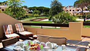 Un balcón con mobiliario exterior, una mesa lista para el desayuno y vistas a una piscina y edificios al fondo.