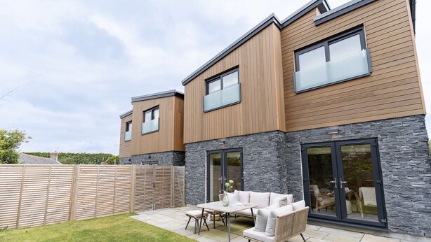 Une maison moderne à deux étages, dotée d’une façade en pierre, d’un revêtement extérieur en bois et d’un patio aménagé avec du mobilier d’extérieur.