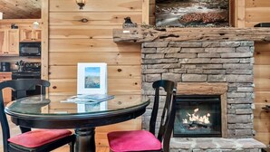 A cozy cabin interior with a stone fireplace, wooden walls, and a dining area with a glass table and chairs.