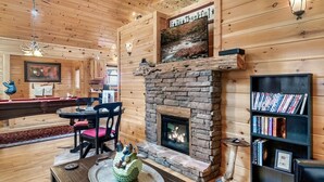 A cozy cabin interior with a stone fireplace, wooden walls, a dining area, and a bookshelf.