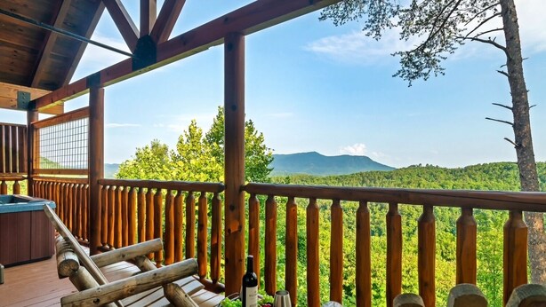 A wooden deck with a hot tub, a table with a bottle of wine and grapes, and a scenic view of mountains and trees.