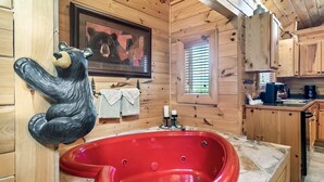 A wooden cabin interior with a red bathtub, a bear sculpture, a framed bear picture, and a window with blinds.