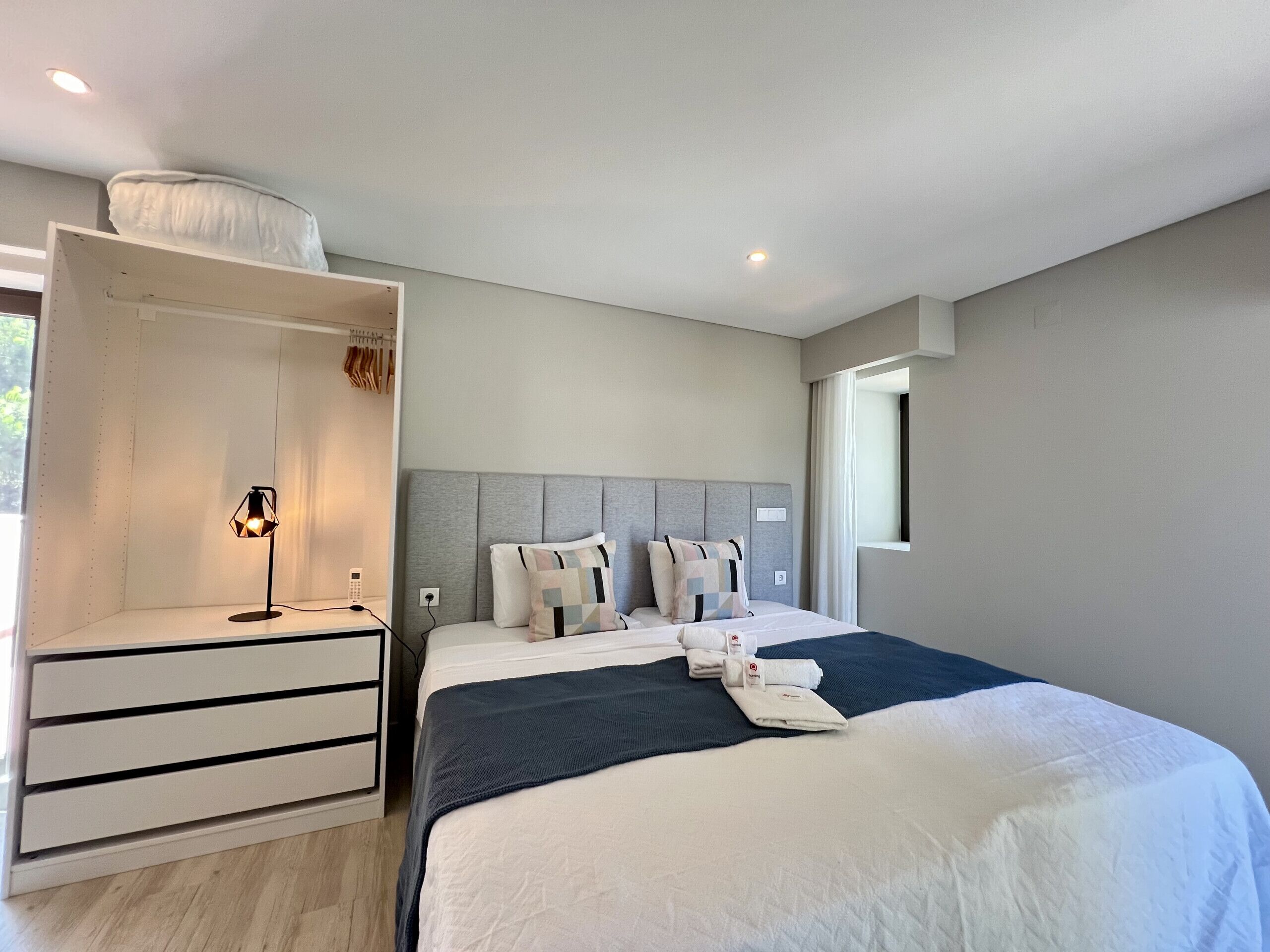 A modern hotel room with a large bed, a built-in wardrobe, a bedside table with a lamp, and a window providing natural light.