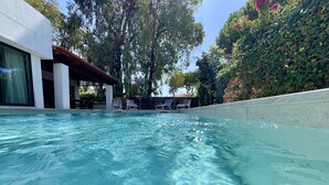 Une piscine aux eaux cristallines, dotée de chaises longues et d’un espace abrité, le tout entouré de verdure et d’un bâtiment.
