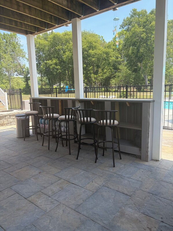An outdoor patio with a bar area, stone flooring, and a pool in the background.