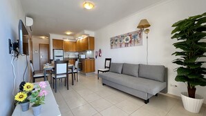 A modern living room with a grey sofa, a dining area with chairs and a table, a kitchen with wooden cabinets, and a large potted plant.