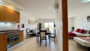 A modern kitchen with wooden cabinets and a built-in oven, a dining area with a table and chairs, and a living room with a sofa and a bed.