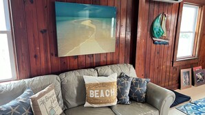Una habitación con paneles de madera, un sofá de cuero color beige, un cojín con motivos playeros y un cuadro grande con una escena de playa.