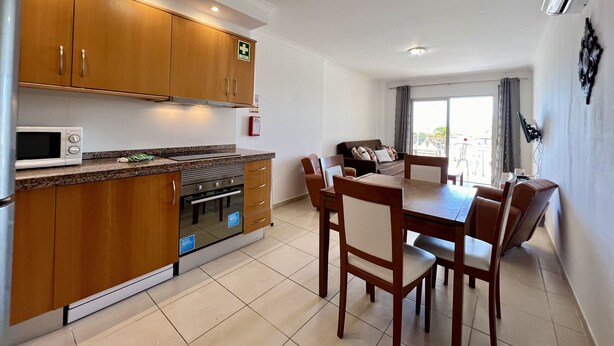 A modern kitchen with wooden cabinets, a built-in oven, and a microwave. There is a dining area with a table and chairs. - Monte Gordo Beach Ocean View 5 by Homing (Vila Real Santo Antonio)