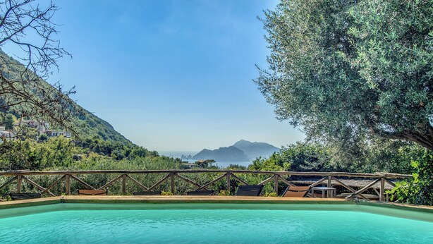 A clear blue pool with a wooden deck and lounge chairs, surrounded by lush greenery and overlooking a mountainous landscape.