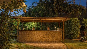 A stone bar with a thatched roof, surrounded by lush greenery and a stone pathway.