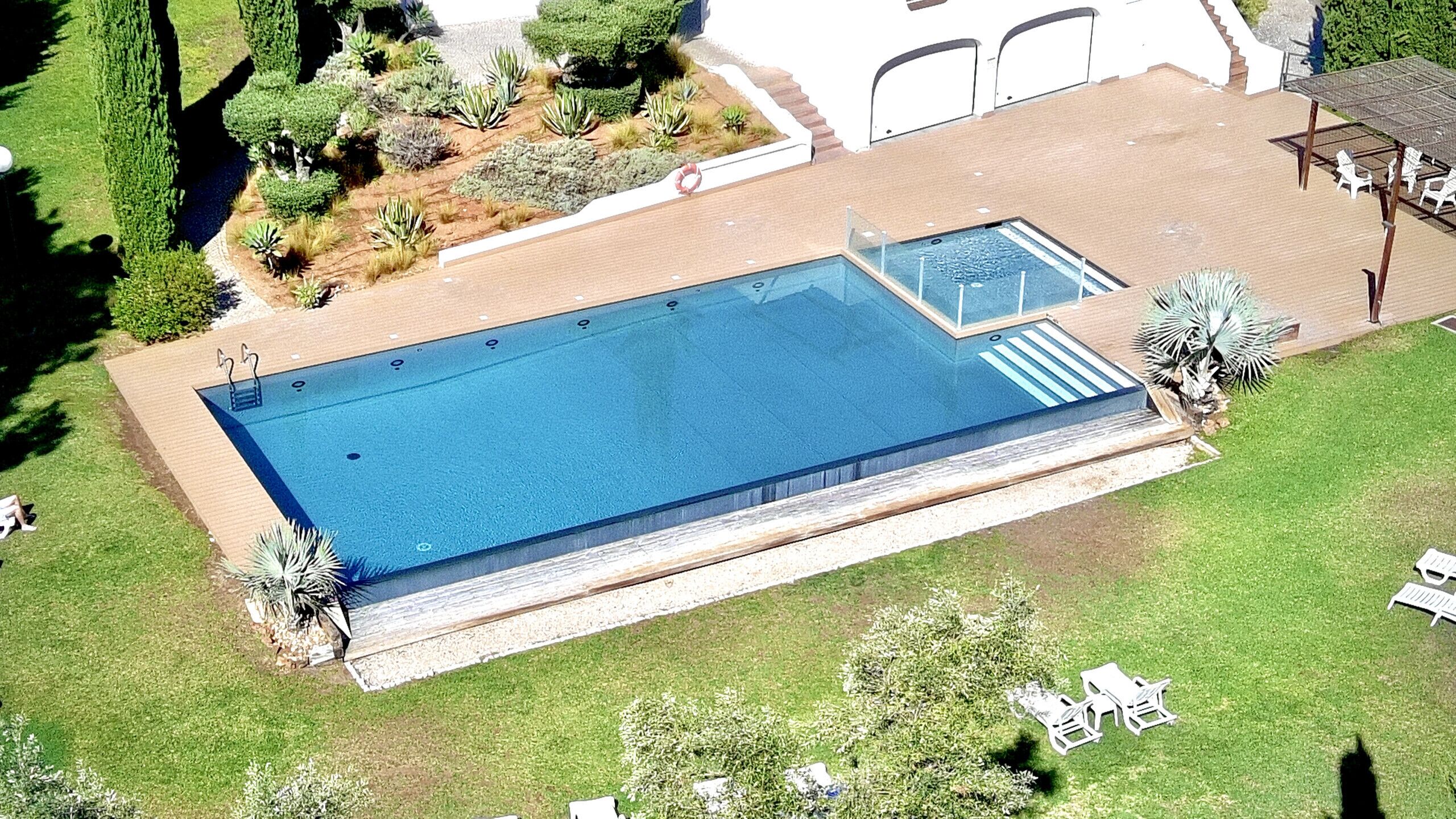 Vista aérea de una piscina rectangular con un pequeño jacuzzi contiguo, rodeada de un césped cuidado y diversas plantas.