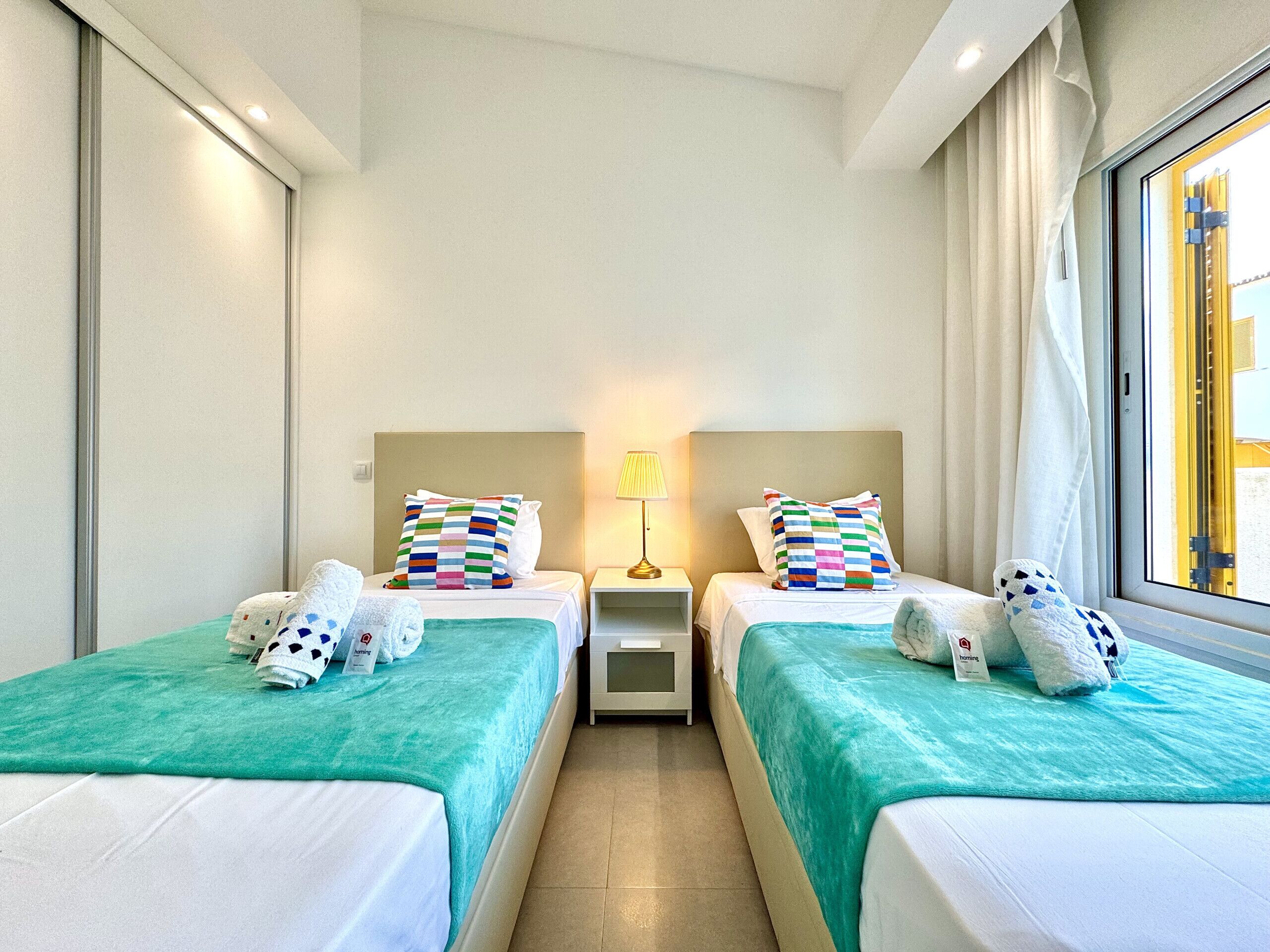 Une chambre d’hôtel avec deux lits, chacun recouvert d’une couvre-lit turquoise et agrémenté de coussins colorés. Une table de chevet avec une lampe se trouve entre les deux lits.