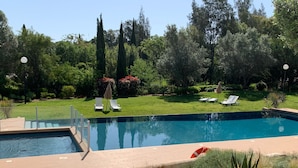A rectangular swimming pool with a clear blue water surface, surrounded by a brick deck. There are two white lounge chairs with umbrellas, and a small red lifebuoy near the pool's edge. The pool is set in a landscaped garden with various plants and trees in the background. - Vilamoura Club Villa 3 by Homing (Loulé)