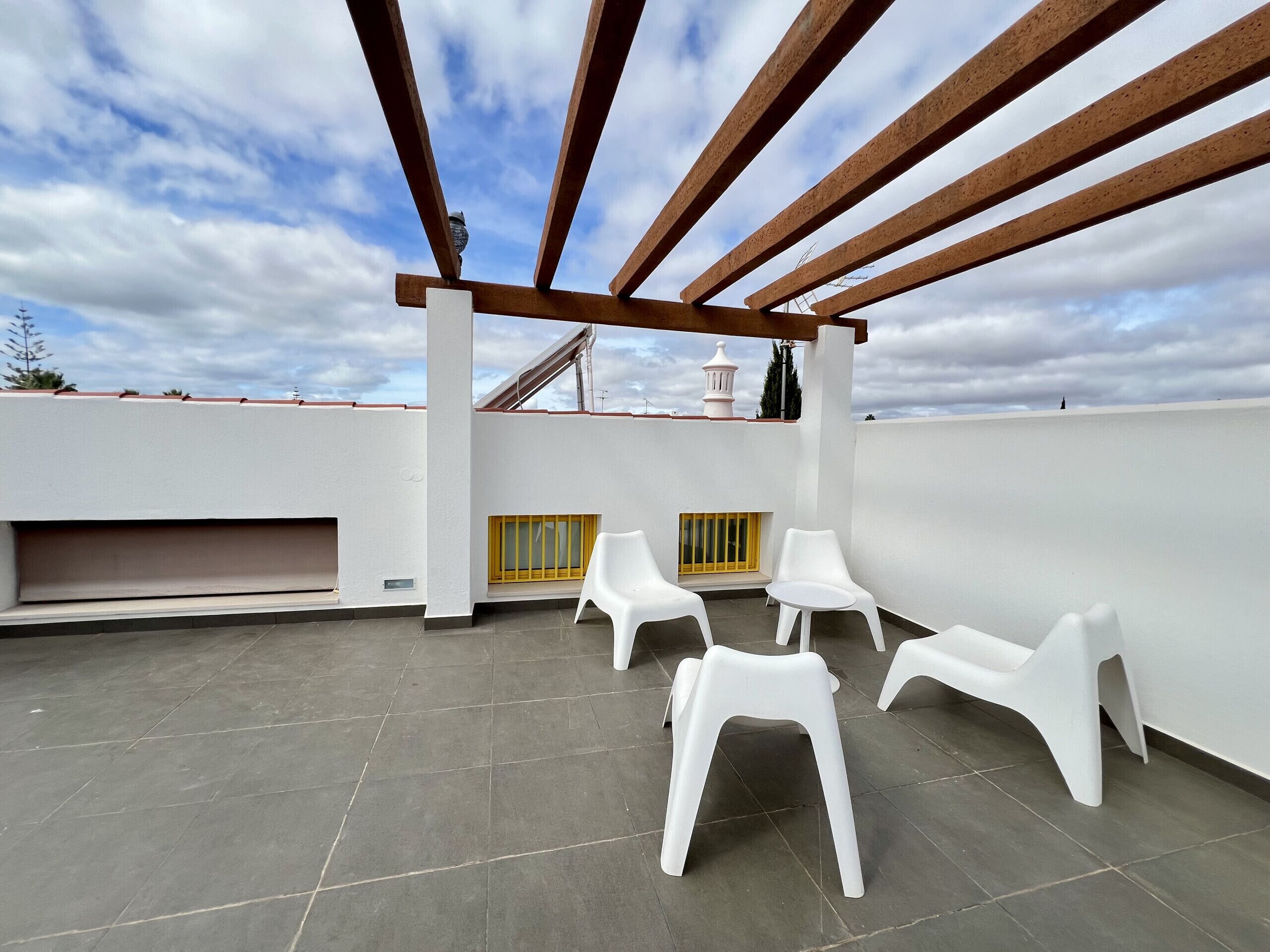A rooftop terrace with white plastic chairs, a pergola, and a view of the sky.