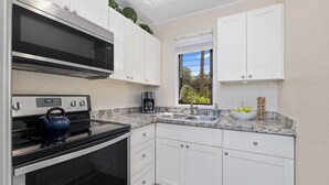 A modern kitchen with white cabinets, a stainless steel oven, a microwave, and a window with a view of greenery. - Kona Bali Kai#164 (Kailua-Kona)