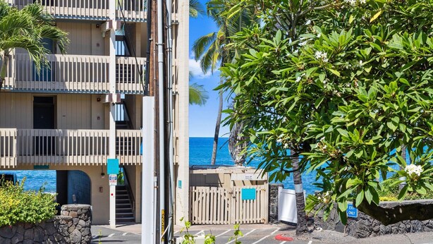 A multi-story building with balconies, a parking area, and a view of the ocean. - Kona Bali Kai#164 (Kailua-Kona)