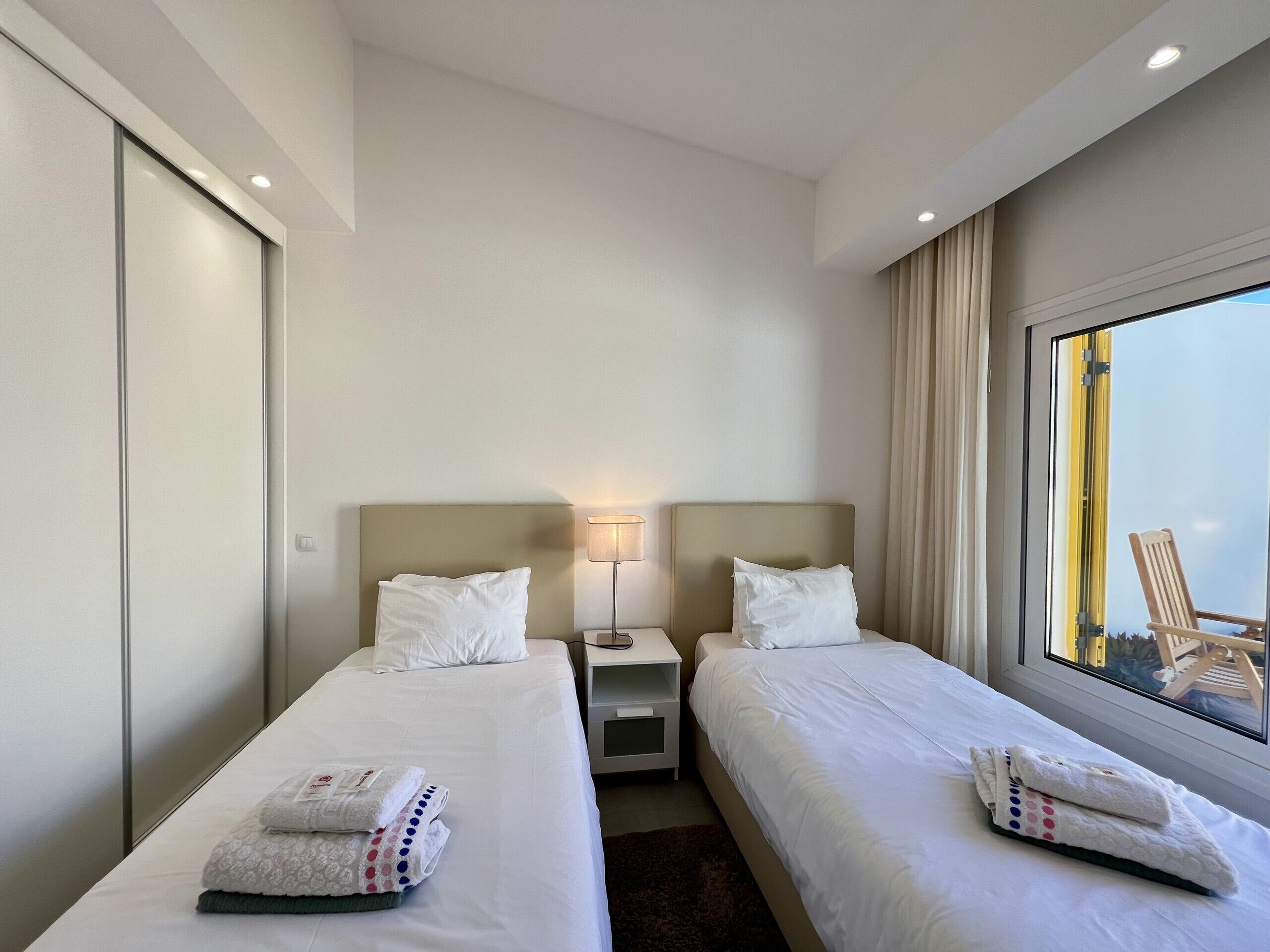 A hotel room with two beds, a nightstand, a lamp, and a window with a view of the outside.