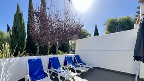 Zona relax all'aperto illuminata dal sole, con poltrone bianche e cuscini blu, circondata da alberi e un muro bianco.