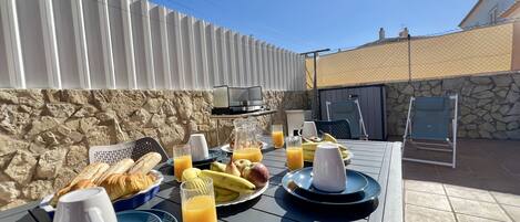 An outdoor dining area with a stone wall, a grill, and a table set with breakfast items including bread, fruits, and drinks.