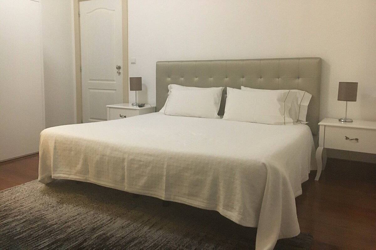 Una cama bien hecha con cabecera gris, dos mesitas de noche con lámparas y una puerta a la izquierda.