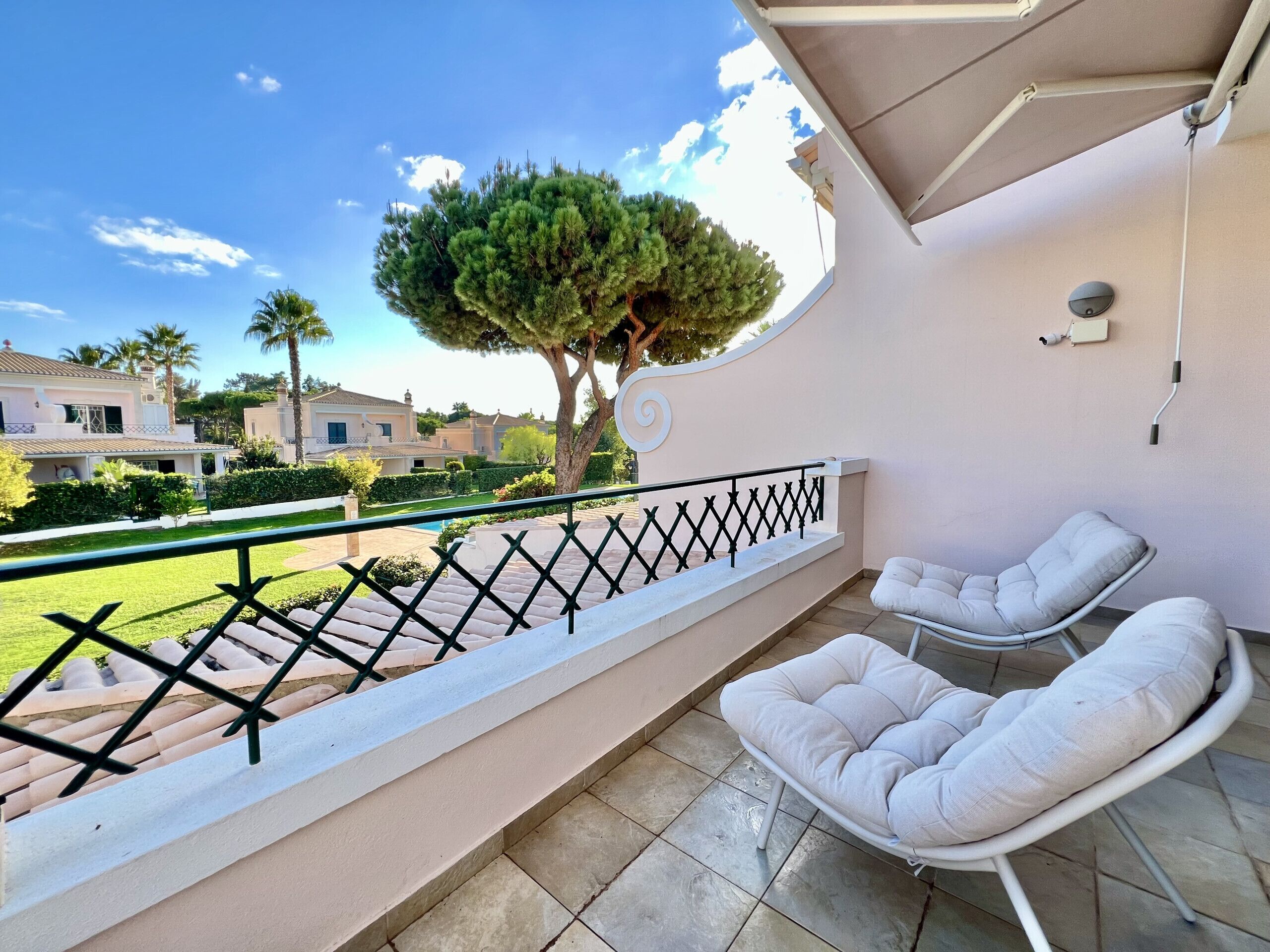 Un balcón con una silla blanca acolchada y una mesita pequeña, con vistas a un jardín cuidado y casas vecinas.